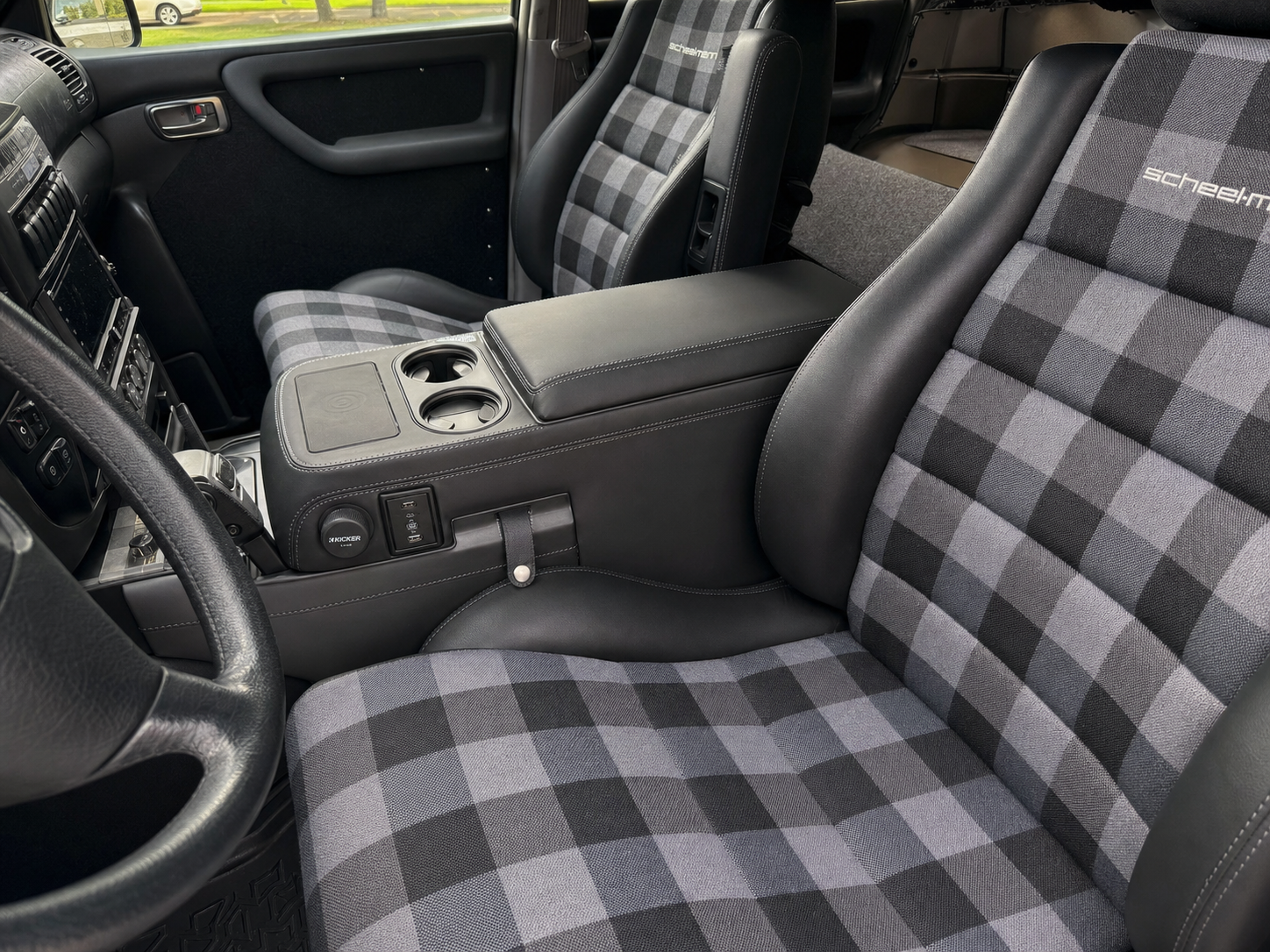 Custom center console wrapped in black leather with grey contrast stitching matching the Scheelmann seats, wireless charging pad, cup holders, Kicker subwoofer knob, USB-C ports, leather strap storage cubby